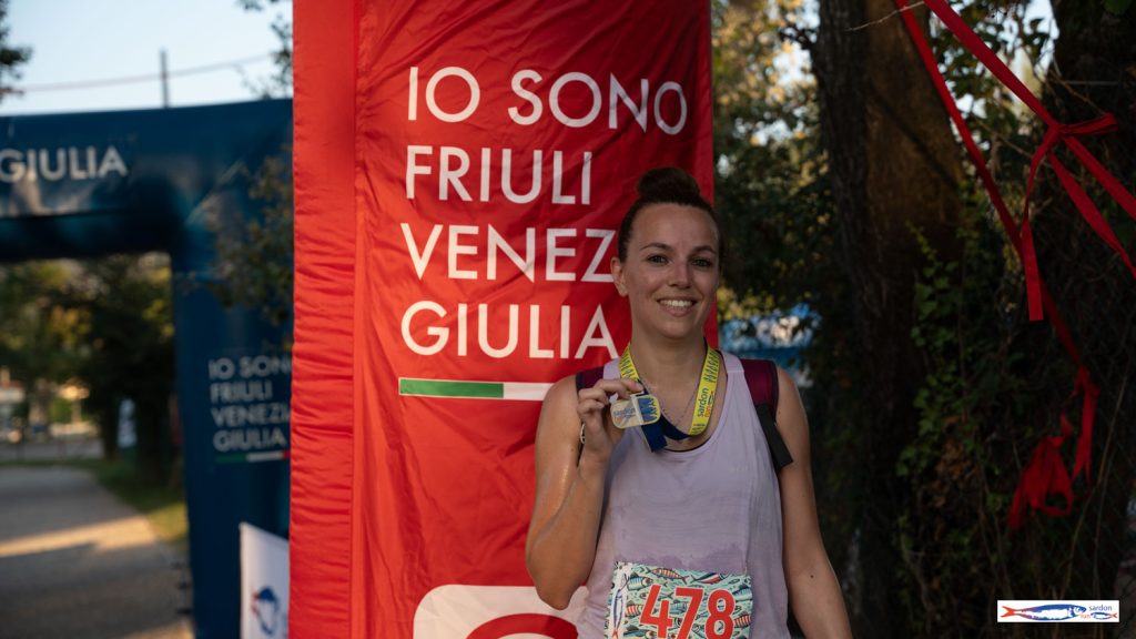Una finisher mostra la medaglia della Sardon Run davanti al totem IO SONO Friuli Venezia Giulia