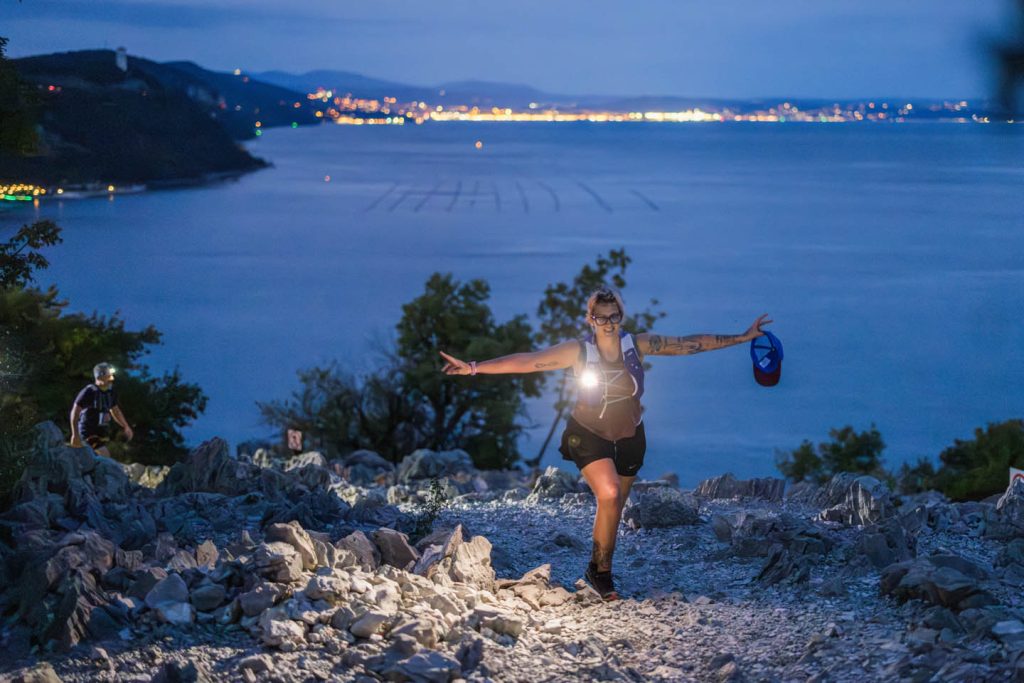 Trail runner con lampada frontale sulla pietraia carsica al crepuscolo, con le luci del Golfo di Trieste sullo sfondo — Sardon Run